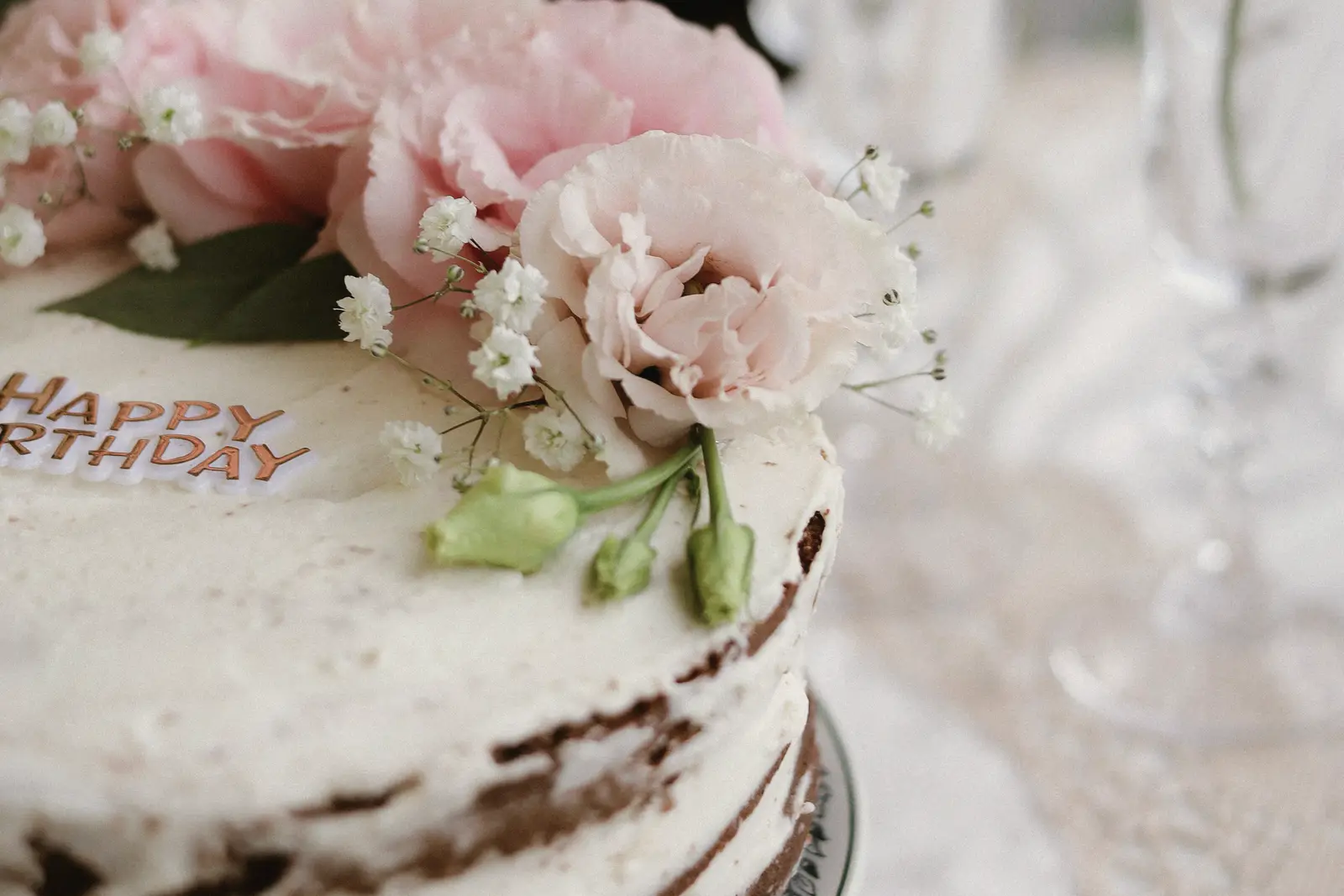 Stock photo: a semi-naked birthday cake topped with pink peonies and baby’s breath, beside crystal glasses