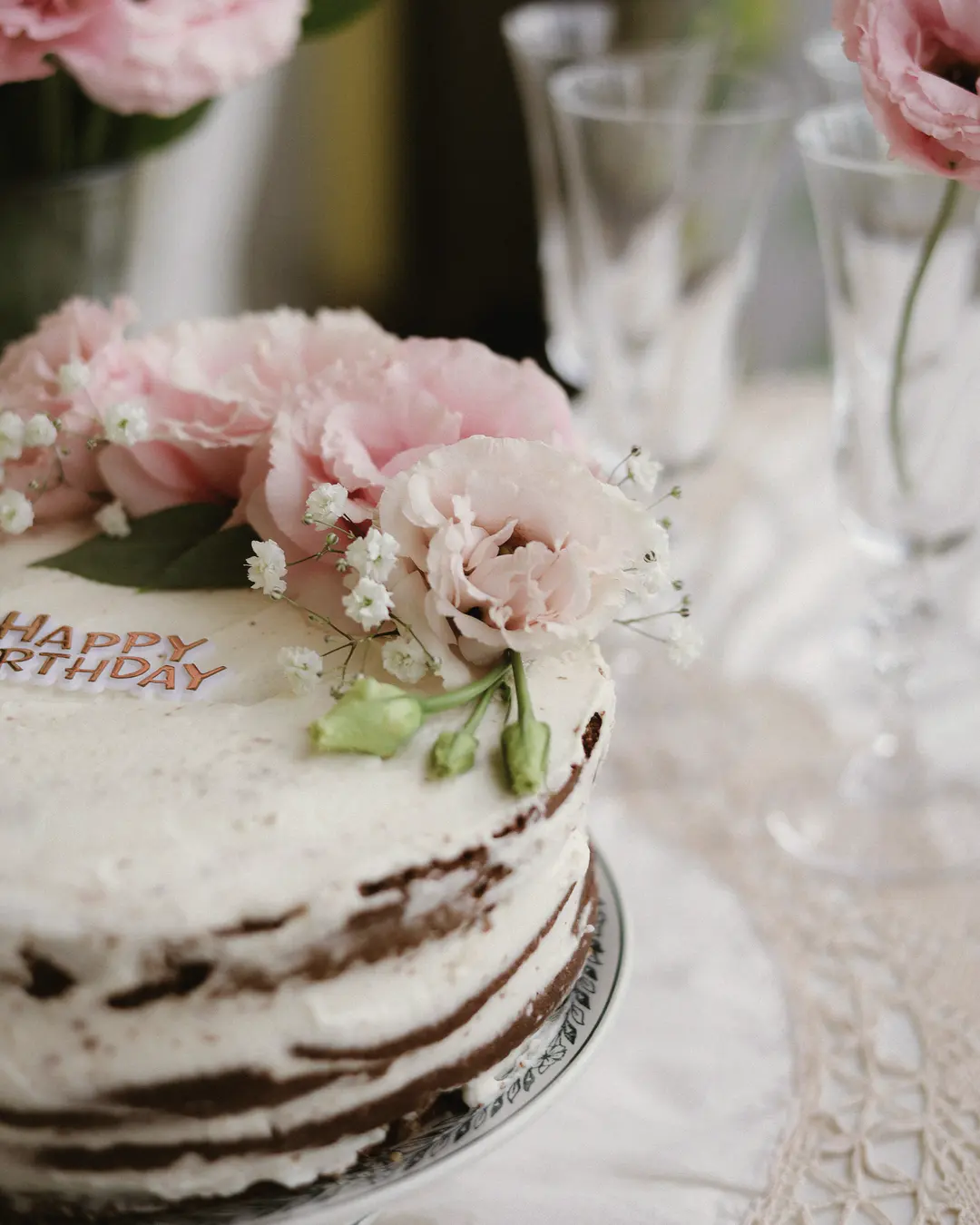 Stock photo: a semi-naked birthday cake topped with pink peonies and baby’s breath, beside crystal glasses