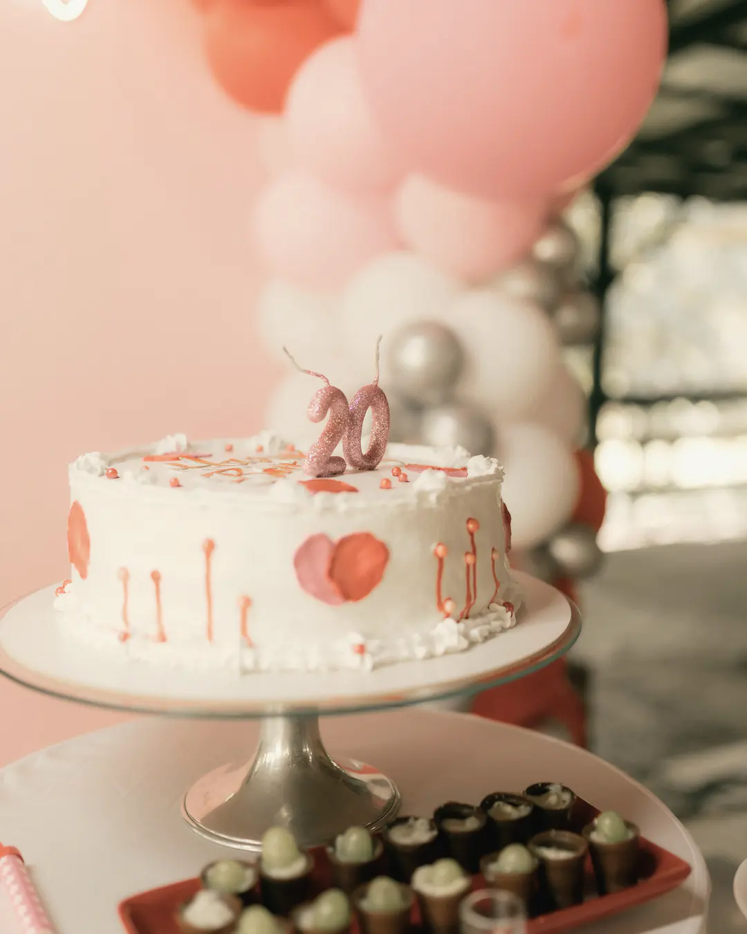 Stock photo: a white birthday cake with pink details and a “20” topper on a cake stand, in front of pink balloons