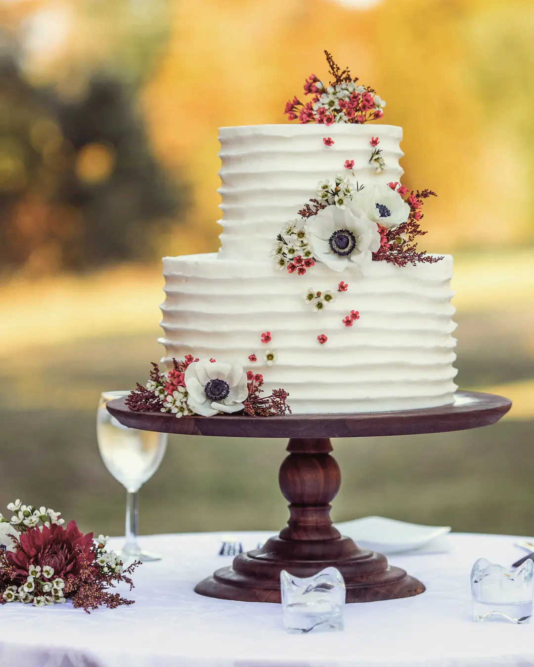 Stock photo: a white combed-buttercream tiered cake with red and white flowers on a wooden stand, outdoors in golden light