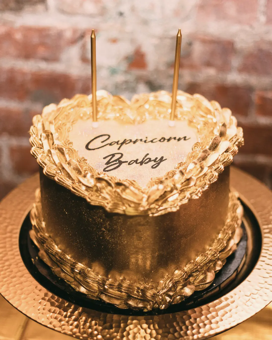 Stock photo: a gold vintage-style heart cake with ruffled piped borders and the message “Capricorn Baby”, on a hammered gold cake board