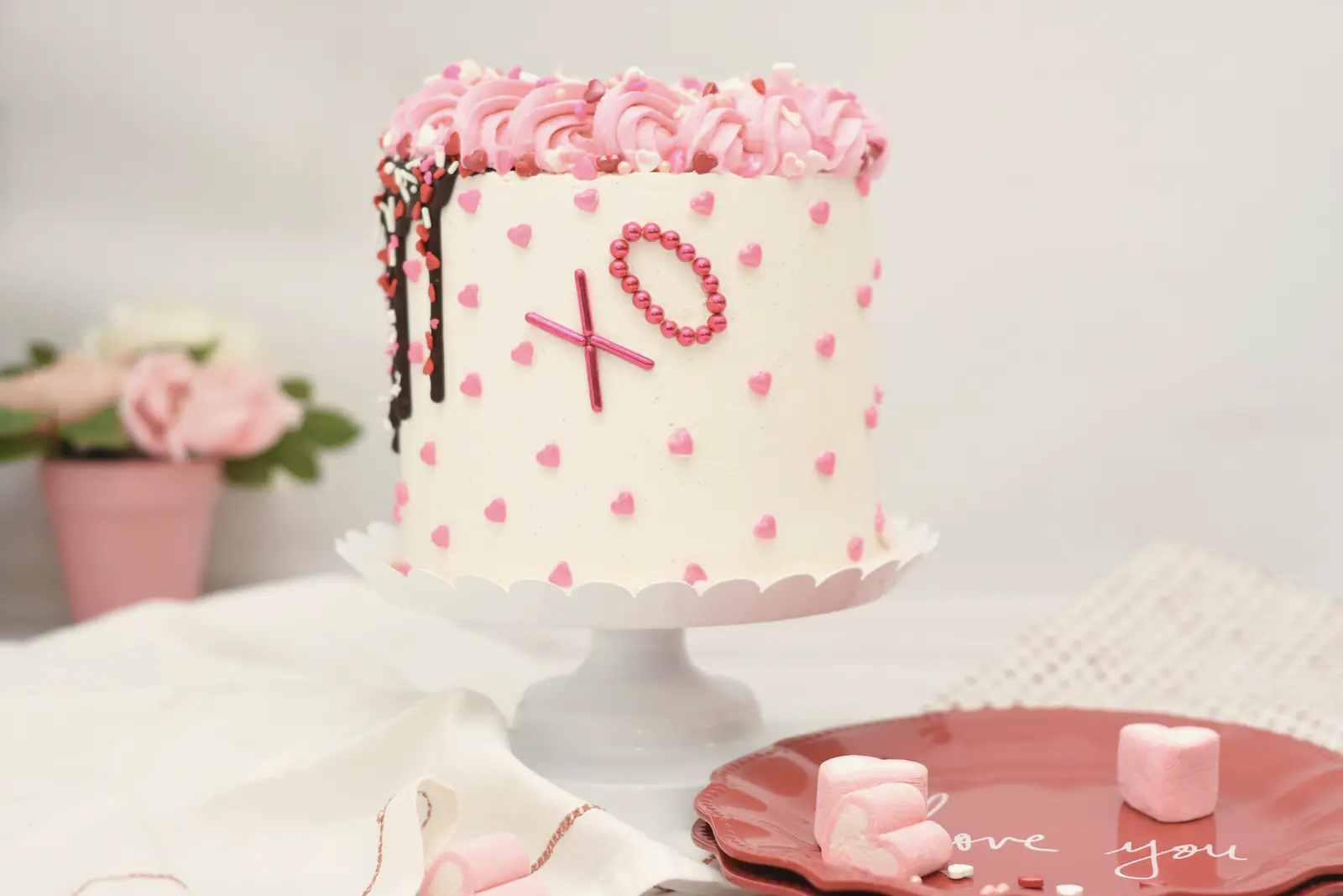 Stock photo: a tall white cake covered in tiny piped pink hearts, pink rosettes and an “XO” design, on a white cake stand