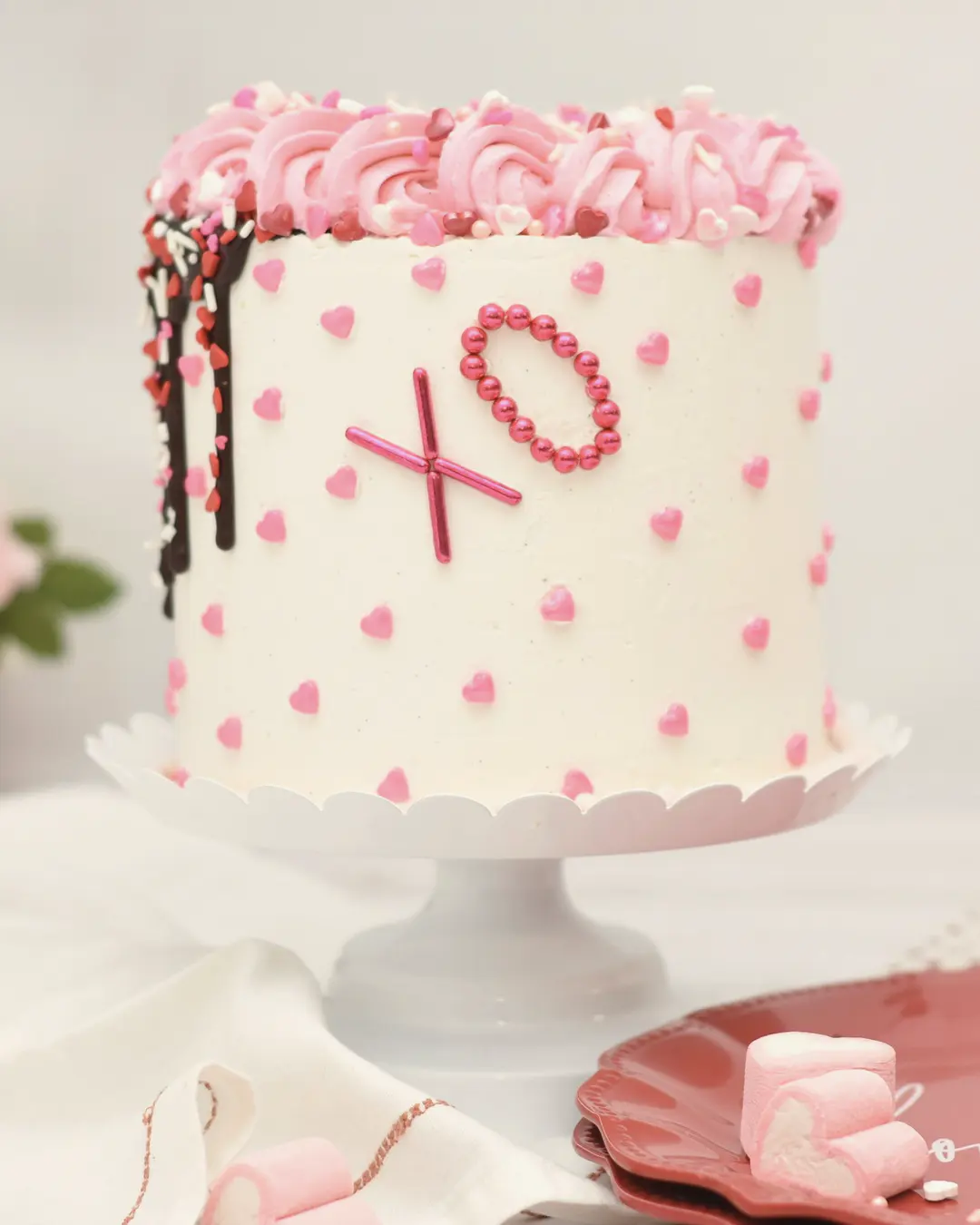 Stock photo: a tall white cake covered in tiny piped pink hearts, pink rosettes and an “XO” design, on a white cake stand